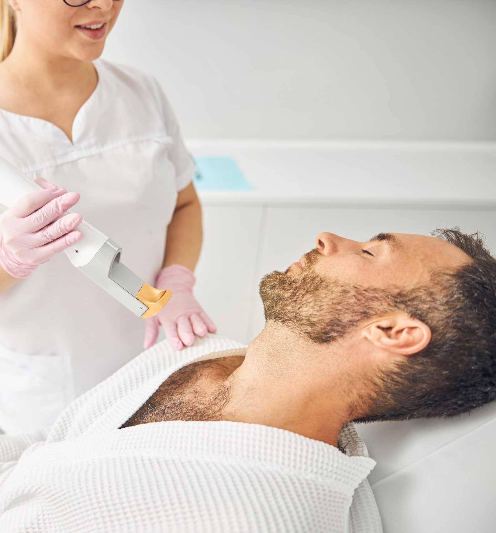 Charming female esthetician holding laser hair removal device and smiling while bearded young man lying on daybed