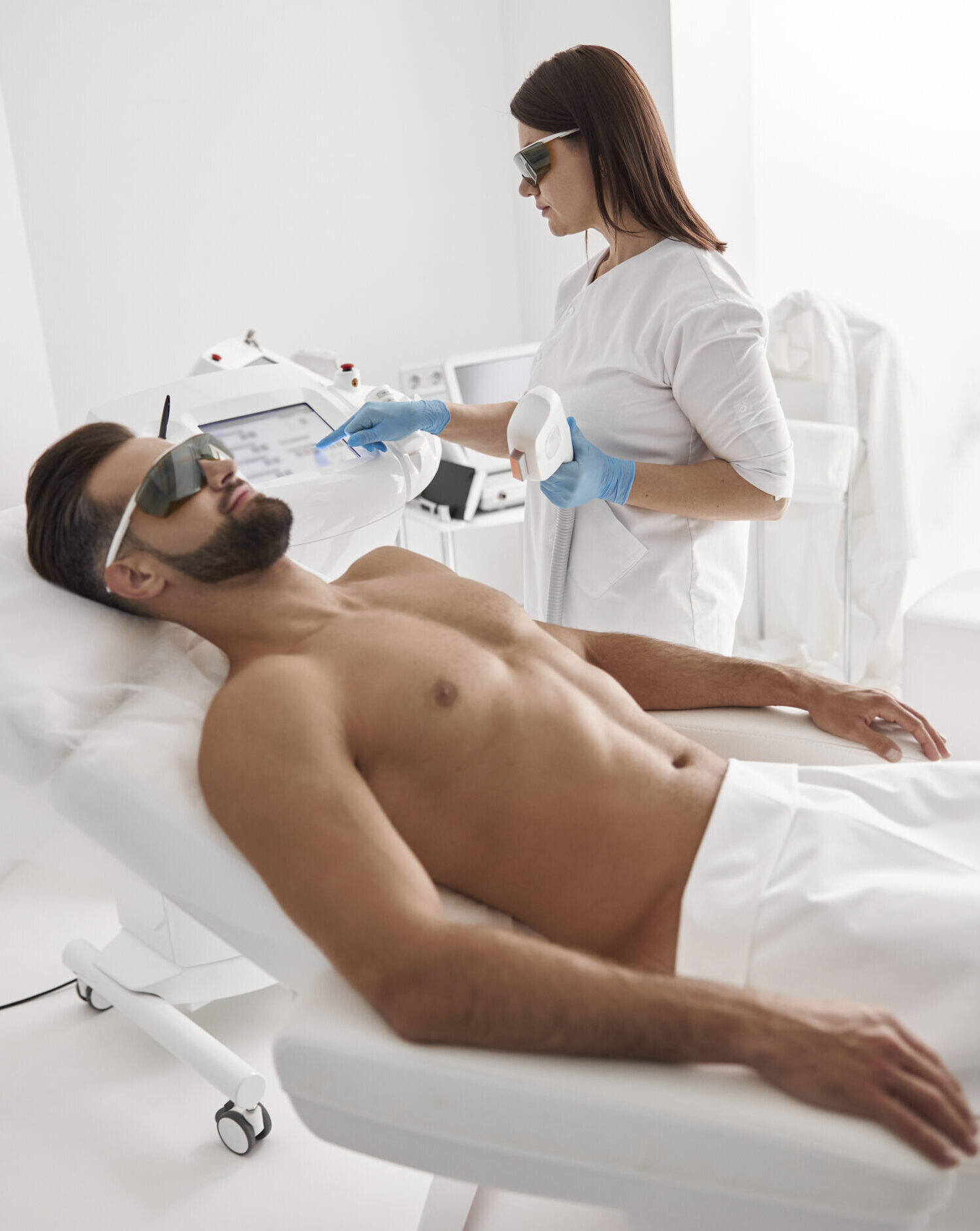 Brunette lady master in white uniform tunes contemporary equipment for laser epilation while mature man client lies on couch in office of beauty clinic