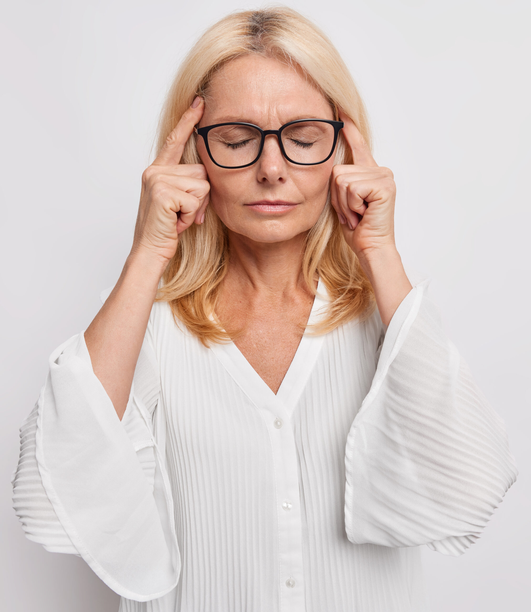 Middle aged blonde attractive woman keeps fingers on temples suffers from headache tries to remember something important stands with closed eyes wears spectacles white blouse faces tough problem